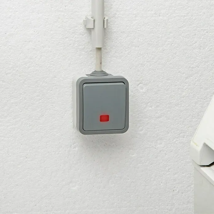 Voltomat Blueline Feuchtraum-Kontrollschalter - Grau, Aufputzmontage, IP54 - Nahaufnahme eines grauen Lichtschalters mit einer kleinen roten Anzeigeleuchte an einer weißen Wand, verbunden mit einem grauen Kabelkanal.