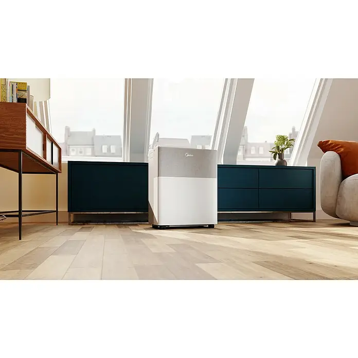 Midea Klimasplitgerät PortaSplit Cool 8.000 Btu - Kühlleistung: 2.300 W, Raumgröße: 28 m² - Tragbares Midea-Gerät mit weißem Sockel und grauem Stoff auf Holzfußboden in hellem Wohnzimmer.