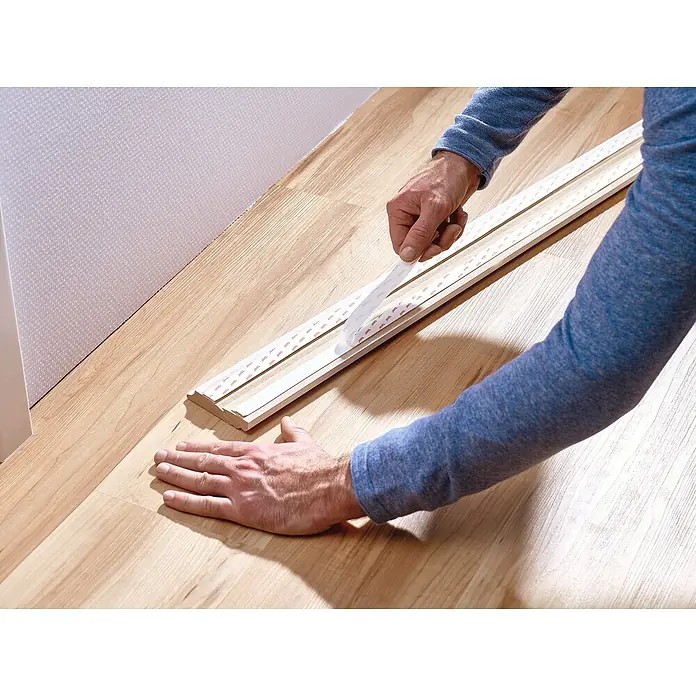 Tesa Powerbond Montageband für Tapete und Putz - L x B: 5 m x 19 mm - Person klebt doppelseitiges tesa Klebeband auf eine weiße Sockelleiste auf einem Holzboden.