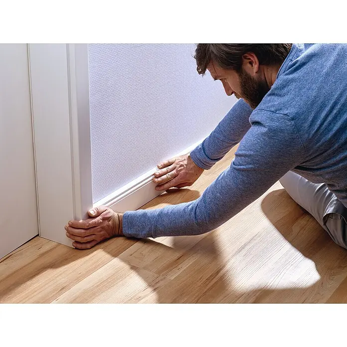 Tesa Powerbond Montageband für Tapete und Putz - L x B: 5 m x 19 mm - Mann montiert weiße Sockelleisten an einer Wand neben einem Türrahmen und einem Holzfußboden.