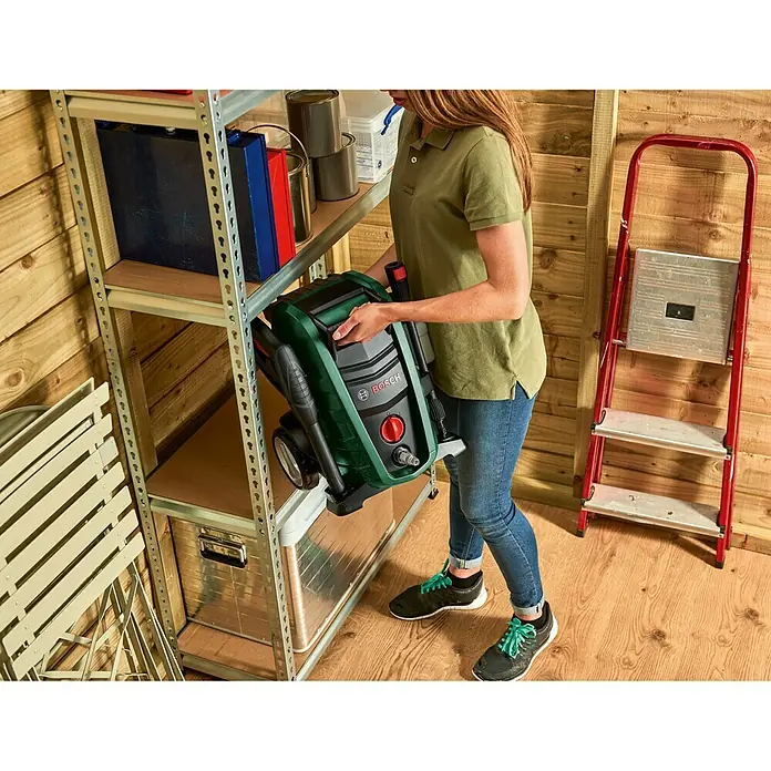 Bosch Hochdruckreiniger UniversalAquatak 130 - 1.700 W, Fördermenge: 380 l/h - Eine Frau hält einen tragbaren Bosch-Hochdruckreiniger in einem Schuppen mit Regalen und einer Leiter.