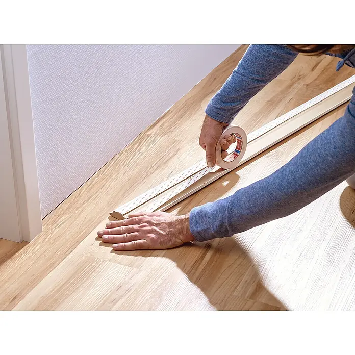 Tesa Powerbond Montageband für Tapete und Putz - L x B: 5 m x 19 mm - Person bringt doppelseitiges Klebeband auf die Rückseite einer hellen Fußleiste auf Holzfußboden an, zur Montagevorbereitung.