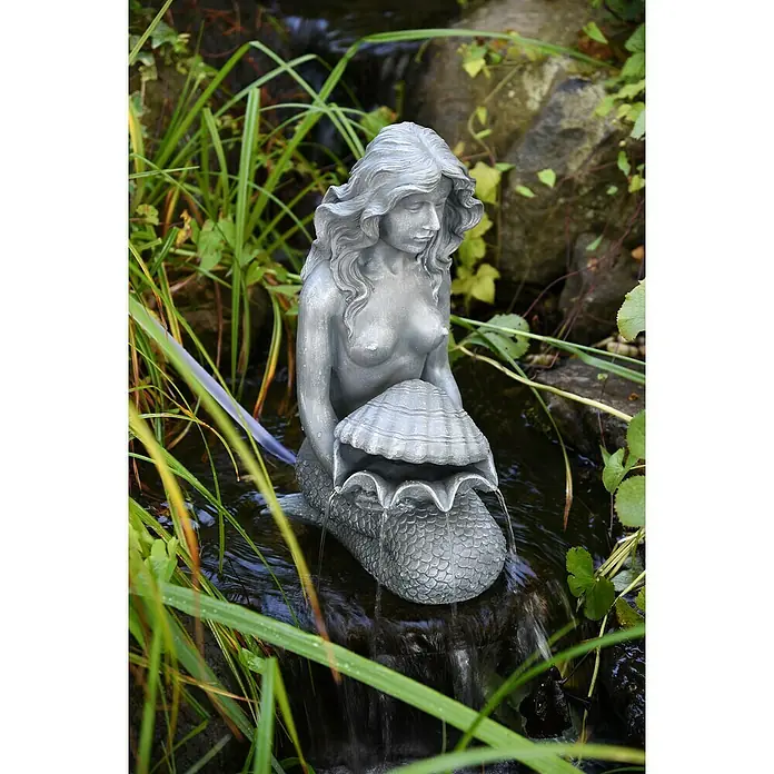 Heissner Wasserspeier Meerjungfrau mit Muschel - L x B x H: 200 x 300 x 460 mm - Graue Meerjungfrauen-Brunnenfigur mit Wasserfluss aus einer Muschel, inmitten grüner Pflanzen im Teich.