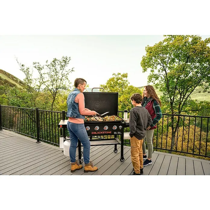 Blackstone Plancha Grill 28" - Superficie de la parrilla (An x Pr): 73 x 46 cm, Negro, Número de quemadores: 2 ud. - Una mujer prepara comida en una parrilla Blackstone mientras dos niños observan. La parrilla se encuentra en una terraza con vistas a un entorno natural.