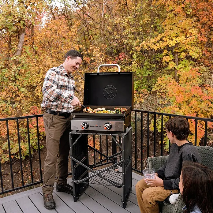 Blackstone Plancha Grill 22" - Superficie de la parrilla (An x Pr): 57 x 41,5 cm, Negro, Número de quemadores: 2 ud. - Hombre asando a la parrilla en una plancha Blackstone al aire libre mientras los niños miran. La plancha Blackstone es portátil.