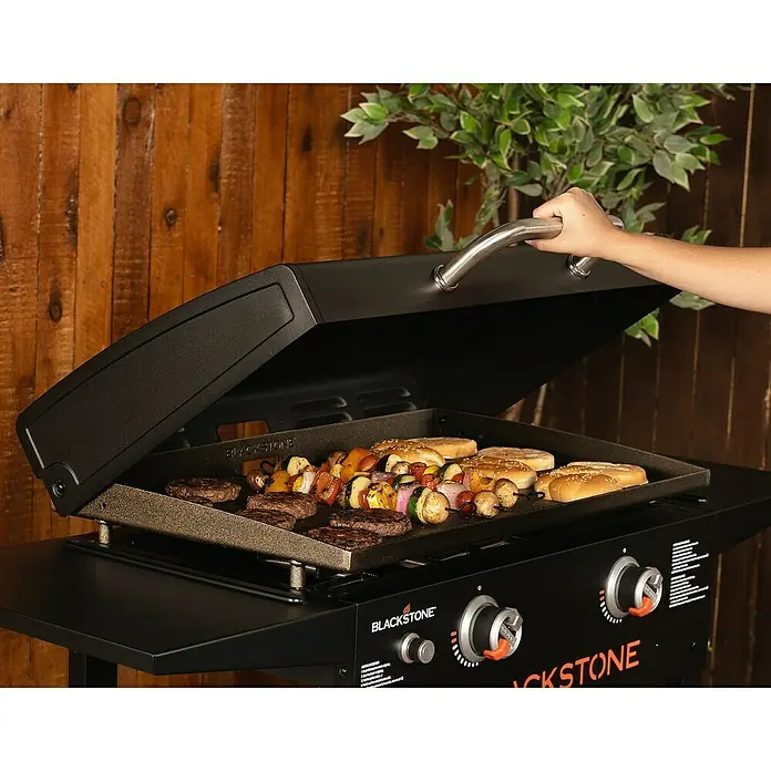 Blackstone Plancha Grill 28" - Superficie de la parrilla (An x Pr): 73 x 46 cm, Negro, Número de quemadores: 2 ud. - Parrilla Blackstone con hamburguesas, brochetas de verduras y panes bajo la tapa abierta.