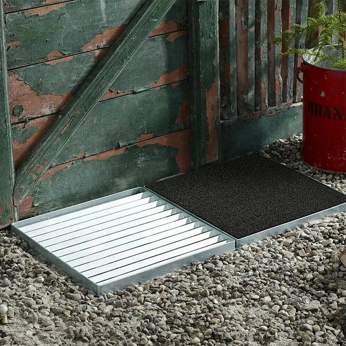 Plus Fußabtreter Cubic - 40 x 80 x 3,5 cm, Schwarz, Silber, Ausführungseigenschaft: Kokosmatte seitlich - Fußabtreter-Set mit einem Gitterrost und einer Schmutzfangmatte in Metallrahmen auf Kies vor einer Holzwand.