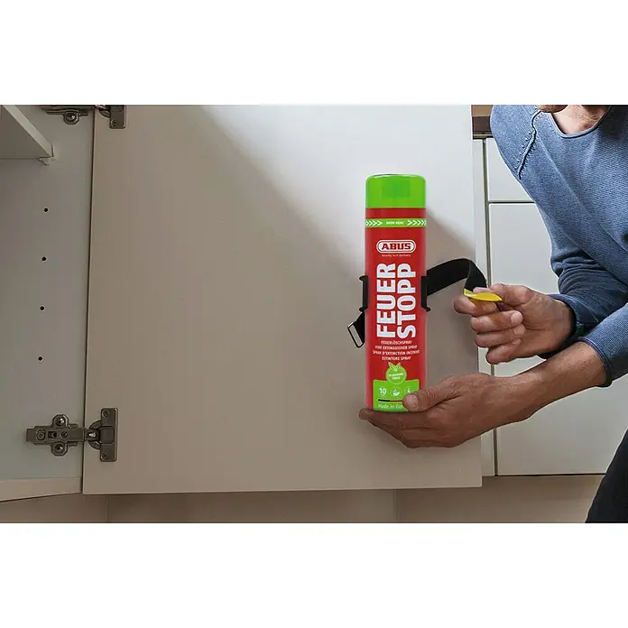 Abus Feuerlöschspray AFS625 - Geeignet für: Elektrische Anlagen bis 1.000 V (min. Abstand 1 m), 625 ml - Nahaufnahme eines ABUS Feuerstopp-Feuerlöschsprays, das an einer Küchenschranktür befestigt ist und von einer Person in der Hand gehalten wird.