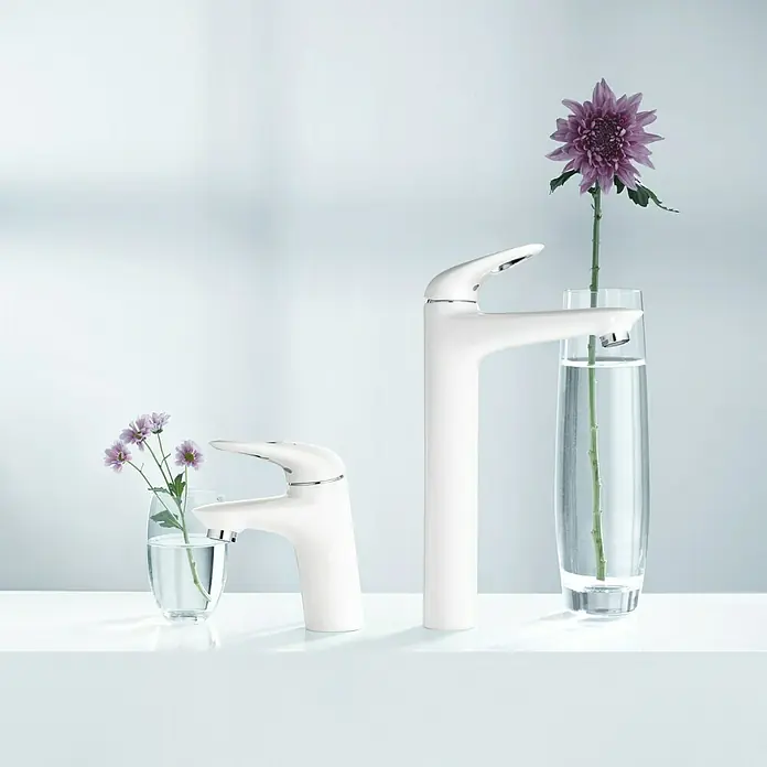 Grohe Eurostyle Waschtischarmatur S-Size - Weiß/Chrom, Glänzend - Zwei moderne, weiße Wasserhähne unterschiedlicher Größe mit Blumen in Gläsern, die eine stilvolle Badezimmeratmosphäre schaffen.