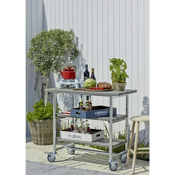 Plus Gartentisch / Pflanztisch Urban - 100 x 50 x 90 cm, Graubraun - Ein Servierwagen aus Metall mit drei Ablagen, auf dem Getränke und Speisen für eine Gartenparty bereitstehen.