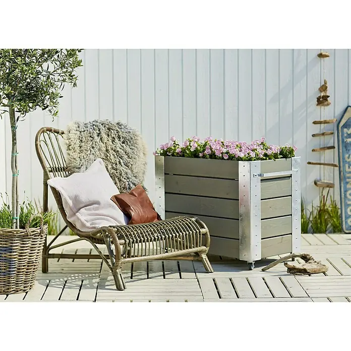 Plus Pflanzkübel eckig Cubic - 87 x 50 x 70 cm, Graubraun, Kiefer - Eine Szene im Freien mit einem grauen Pflanzkasten mit Metallakzenten, der mit rosa Blumen gefüllt ist, neben einem Rattan-Stuhl mit Kissen.