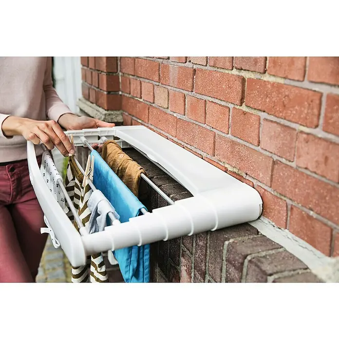 Vileda Wandwäschetrockner Genius 70 - Länge Wäscheleine: 5 m, Farbe: Weiß - Eine Person, die einen weißen, an einer Ziegelmauer montierten Wäscheständer mit darauf hängenden Kleidungsstücken benutzt.