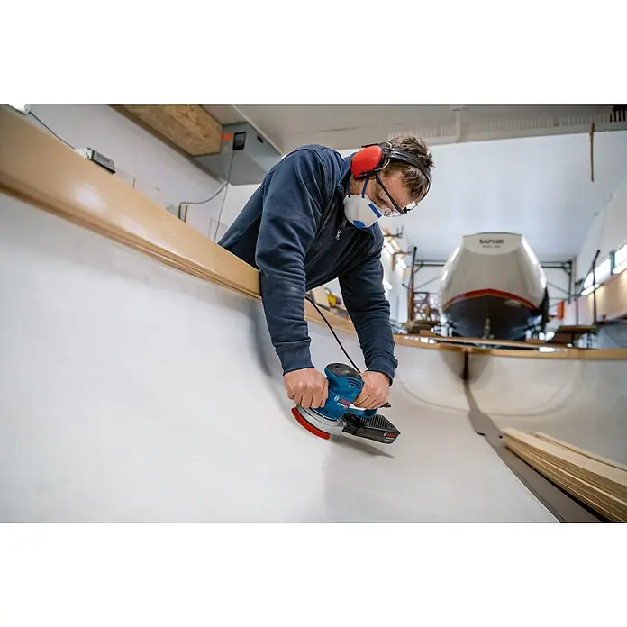 Bosch Professional Elektro-Exzenterschleifer GEX 34-125 - 340 W, Durchmesser Schleifteller: 125 mm, Leerlaufdrehzahl: 4.500 U/min - 12.000 U/min - Ein Handwerker schleift mit einem blauen Bosch-Exzenterschleifer ein Boot, während er Schutzbrille und Ohrenschützer trägt.