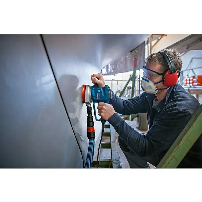 Bosch Professional Elektro-Exzenterschleifer GEX 34-125 - 340 W, Durchmesser Schleifteller: 125 mm, Leerlaufdrehzahl: 4.500 U/min - 12.000 U/min - Ein Mann benutzt einen Bosch Exzenterschleifer an einer Wand, um die Oberfläche zu glätten und vorzubereiten. Schutzausrüstung wird getragen.