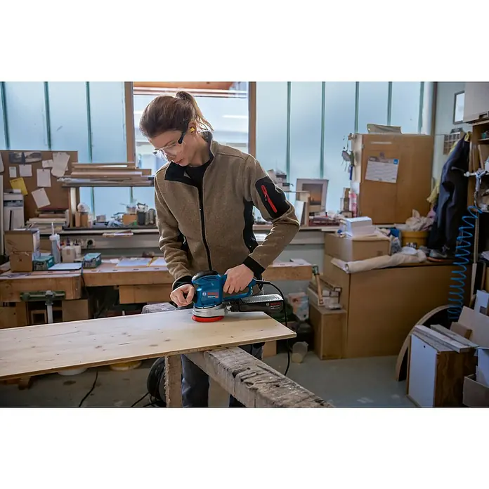 Bosch Professional Elektro-Exzenterschleifer GEX 34-125 - 340 W, Durchmesser Schleifteller: 125 mm, Leerlaufdrehzahl: 4.500 U/min - 12.000 U/min - Frau schleift Holzbrett mit einem Bosch Exzenterschleifer in einer Werkstatt, mit Schutzbrille und Gehörschutz für sicheres Arbeiten.