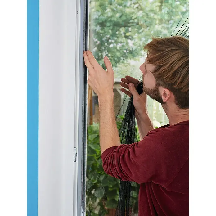 Tesa Insect Stop Mosquitera - An x Al: 65 x 220 cm, Montaje de la mosquitera: Enganche - Un hombre fija una mosquitera magnética a un marco de ventana, asegurando un hogar libre de insectos. La malla negra es fina y el marco es gris.
