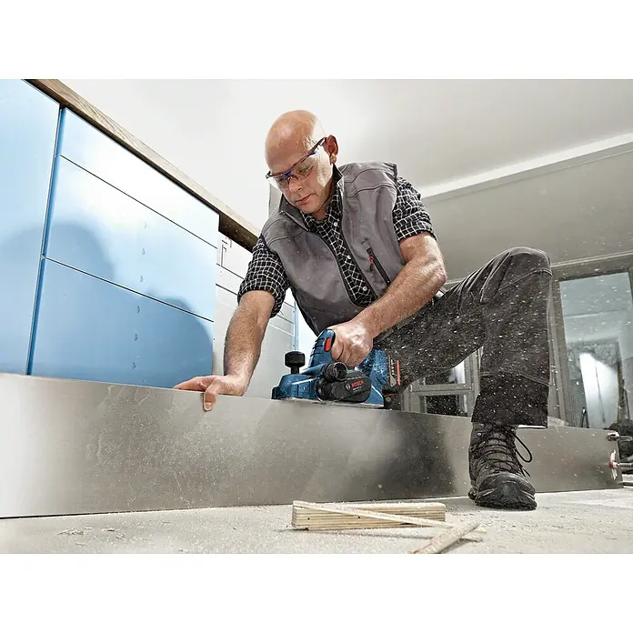 Bosch Professional AMPShare 18V Akku-Hobel GHO 18 V-LI L-Boxx - 18 V, Ohne Akku, Hobeltiefe: 0 mm - 1,6 mm, Leerlaufdrehzahl: 0 U/min - 14.000 U/min - Ein Handwerker benutzt einen Bosch Hobel, um ein Metallstück zu bearbeiten. Das Werkzeug ist ein blauer Akku-Hobel, der präzise Schnitte ermöglicht.