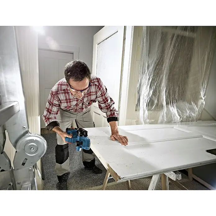 Bosch Professional AMPShare 18V Akku-Hobel GHO 18 V-LI L-Boxx - 18 V, Ohne Akku, Hobeltiefe: 0 mm - 1,6 mm, Leerlaufdrehzahl: 0 U/min - 14.000 U/min - Mann hobelt eine Tür mit einem Bosch Elektrohobel. Ein nützliches Werkzeug für Holzarbeiten, um Oberflächen zu glätten und anzupassen.