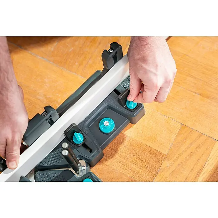 Wolfcraft Gehrungsschneidlade & Schmiege - Kunststoff - Hände befestigen eine weiße Zierleiste in einer schwarz-türkisen Gehrungslade zum Schneiden. Das Werkzeug liegt auf einem Holzfußboden.