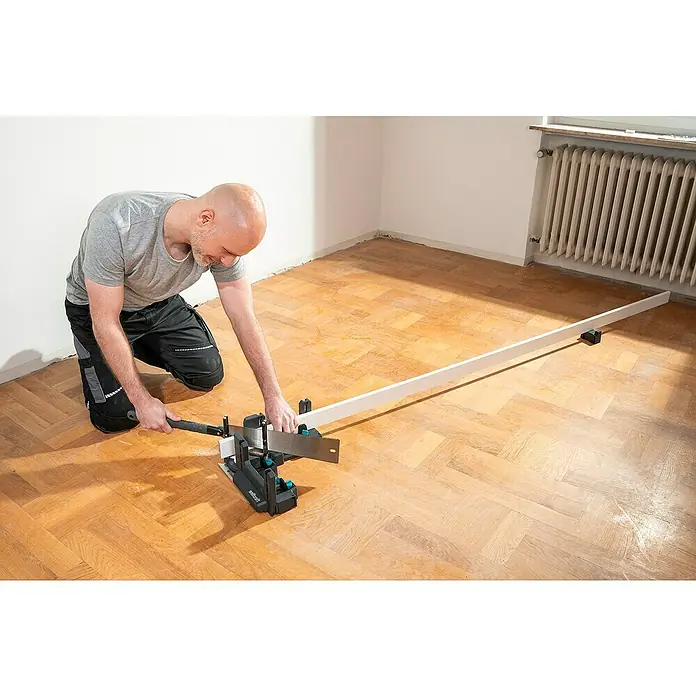 Wolfcraft Gehrungsschneidlade & Schmiege - Kunststoff - Ein Mann kniet und schneidet mit einer Handsäge und einer Schneidlade eine weiße Fußleiste in einem Raum mit Parkettboden.