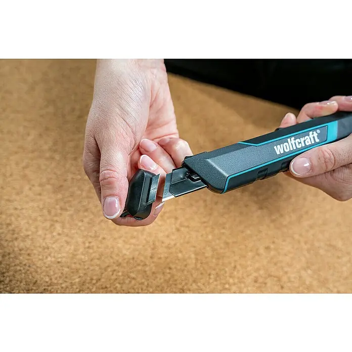 Wolfcraft Cuttermesser Pro Safety - Abbrechklinge, Klingenlänge: 18 mm - Eine Person verwendet den integrierten Klingenabbrecher eines dunkelgrauen Wolfcraft Cuttermessers mit blauen Akzenten, um ein stumpfes Segment zu entfernen.