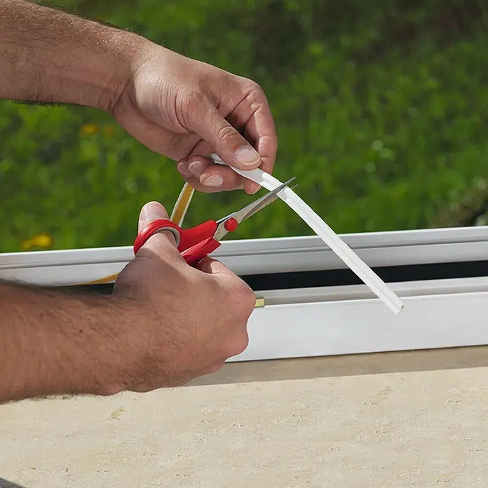 Tesa INSULATION Cinta de sellado - L x An x Al: 6 m x 9 mm x 5,5 mm, Blanco - Un hombre corta una tira de sellado para ventanas o puertas con unas tijeras rojas para un mejor aislamiento.