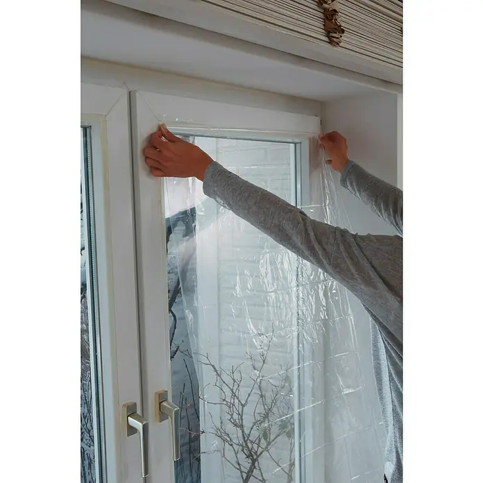 Tesa INSULATION Lámina aislante para ventanas Thermo Cover - 1,5 x 1,7 m, Transparente - Una persona sujeta con ambas manos una película transparente a un marco de ventana blanco para aislar y proteger.