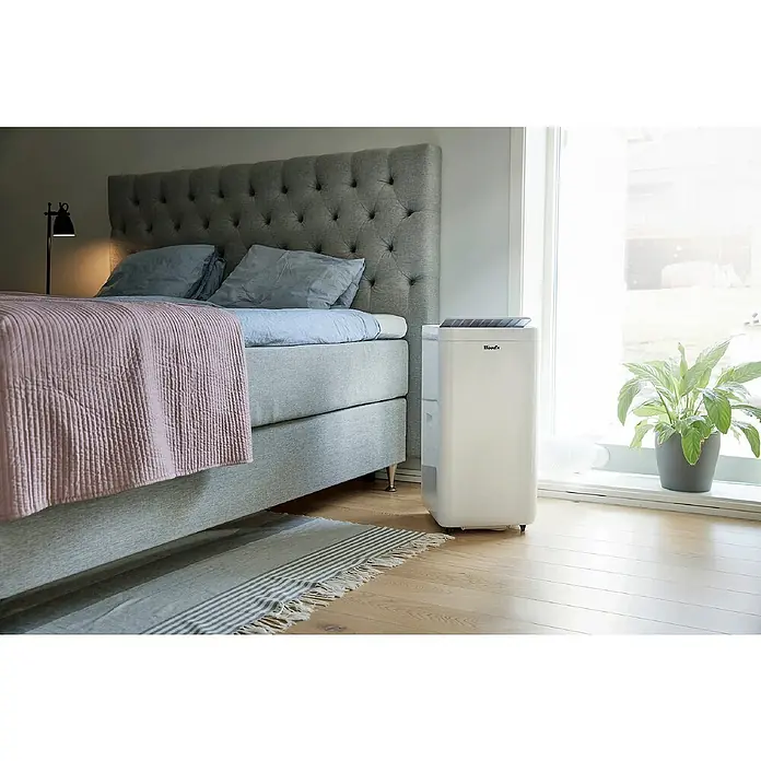 Woods Mobiles Klimagerät Milan 9K WIFI DUO - 2,6 kW, Raumgröße: 26 m², Weiß - Weißer tragbarer Luftentfeuchter/Klimaanlage im Schlafzimmer neben Bett und Fenster.