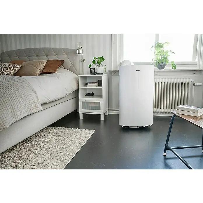 Woods Mobiles Klimagerät Cortina 12K - 3,5 kW, Raumgröße: 35 m², Weiß - Weißes, mobiles Klimagerät in einem modernen Schlafzimmer, neben einem Fenster zur Klimaregelung platziert.