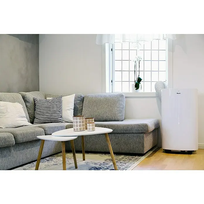 Woods Mobiles Klimagerät Cortina 12K AirSwitch DUO A+ - 3,5 kW, Raumgröße: 35 m², Weiß - Weißes, portables Klimagerät mit Abluftschlauch, neben einem Fenster in einem Wohnzimmer.