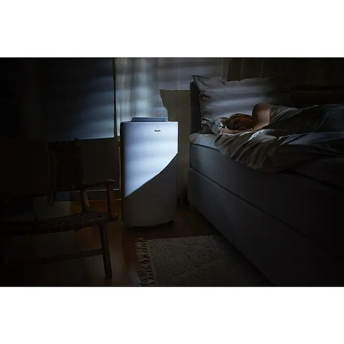 Woods Mobiles Klimagerät Cortina 12K AirSwitch DUO A+ - 3,5 kW, Raumgröße: 35 m², Weiß - Weißes, mobiles Klimagerät/Luftentfeuchter in einem dämmrigen Schlafzimmer neben einer schlafenden Person.