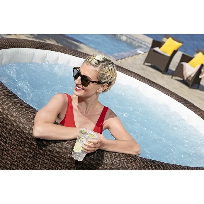 Bestway Lay-Z-Spa Mobil-Whirlpool Bali - Ø x H: 216 x 71 cm, Braun, 1.260 l - Eine Frau entspannt in einem braunen aufblasbaren Whirlpool mit Geflechtmuster, genießt das Wasser und hält ein Getränk.