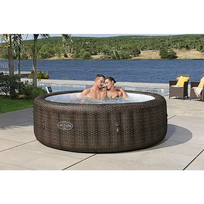 Bestway Lay-Z-Spa Mobil-Whirlpool Bali - Ø x H: 216 x 71 cm, Braun, 1.260 l - Ein Lay-Z-Spa Bali Whirlpool aus Rattan-Optik, in dem ein Paar entspannt, mit Blick auf einen See und üppiges Grün im Hintergrund.