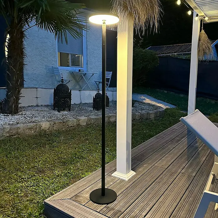 Lavida Akku-Stehleuchte & Tischleuchte Hybrid höhenverstellbar - Schwarz, Warmweiß, 6 W, IP54 - Eine moderne schwarze Stehlampe mit runder, flacher Lichtquelle beleuchtet einen Gartenbereich mit Terrasse und Pflanzen.