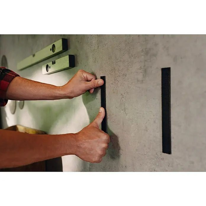 Tesa Ultra Power Klettpad Lock Tape - L x B: 1 m x 25 mm, Schwarz - Nahaufnahme von Händen, die einen Klettstreifen an einer Wand befestigen, neben einer angebrachten Wasserwaage.