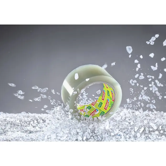 Tesa Pack Paketklebeband Eco & Ultra Strong - Transparent, L x B: 66 m x 50 mm - Nahaufnahme einer Rolle Tesa Eco & Clear Klebeband, umgeben von kleinen weißen Partikeln, die Bewegung suggerieren.
