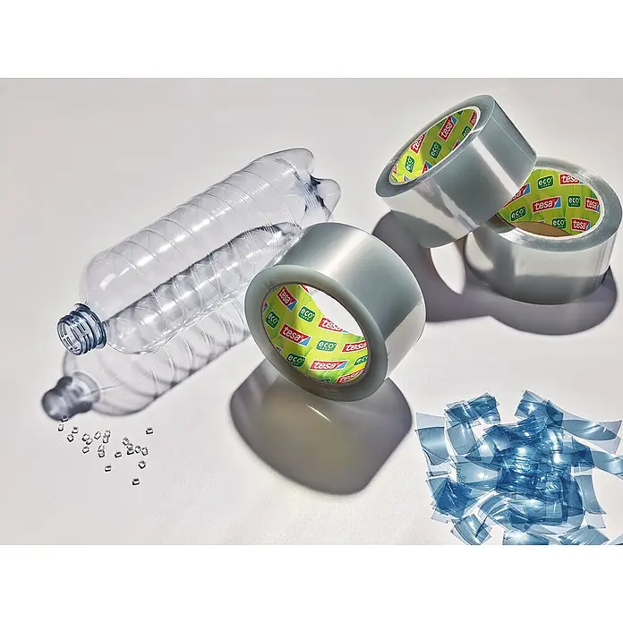 Tesa Pack Paketklebeband Eco & Ultra Strong - Transparent, L x B: 66 m x 50 mm - Ein recycelter PET-Flaschenrohling neben umweltfreundlichem Tesa-Klebeband und zerkleinertem Kunststoff, was das Konzept der Nachhaltigkeit verdeutlicht.
