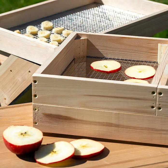 Dobar Dörrschrank Solar - Außenmaß (L x B x H): 102,5 x 52,5 x 98 cm, Holz, Hellbraun - Nahaufnahme von Dörrgeräten aus Holz mit Apfel- und Bananenscheiben auf einem Tisch im Freien, ideal zum Trocknen von Früchten.
