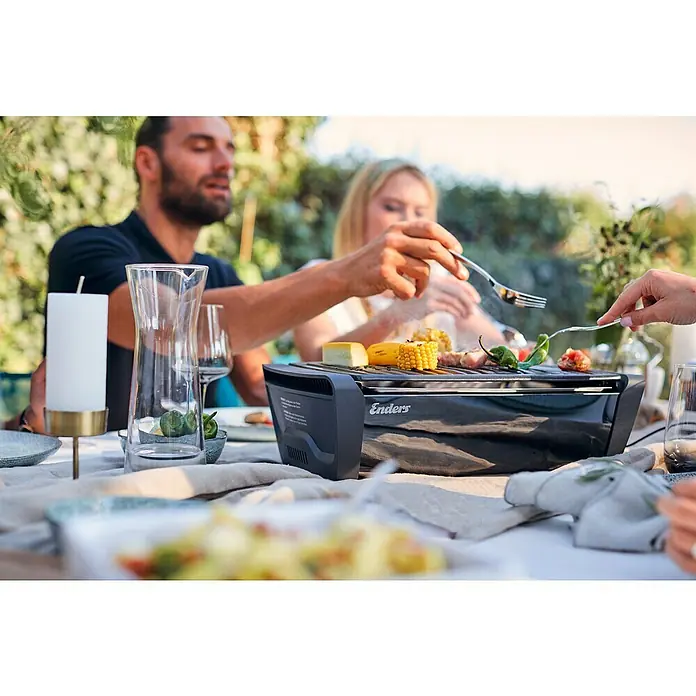 Enders Elektrogrill Aurora - MIrror-Schwarz, Grillfläche (B x T): 37 x 25 cm - Schwarzer Enders Tischgrill auf einem Tisch, darauf brutzeln Mais, Fleisch und Gemüse, umgeben von Menschen im Freien.