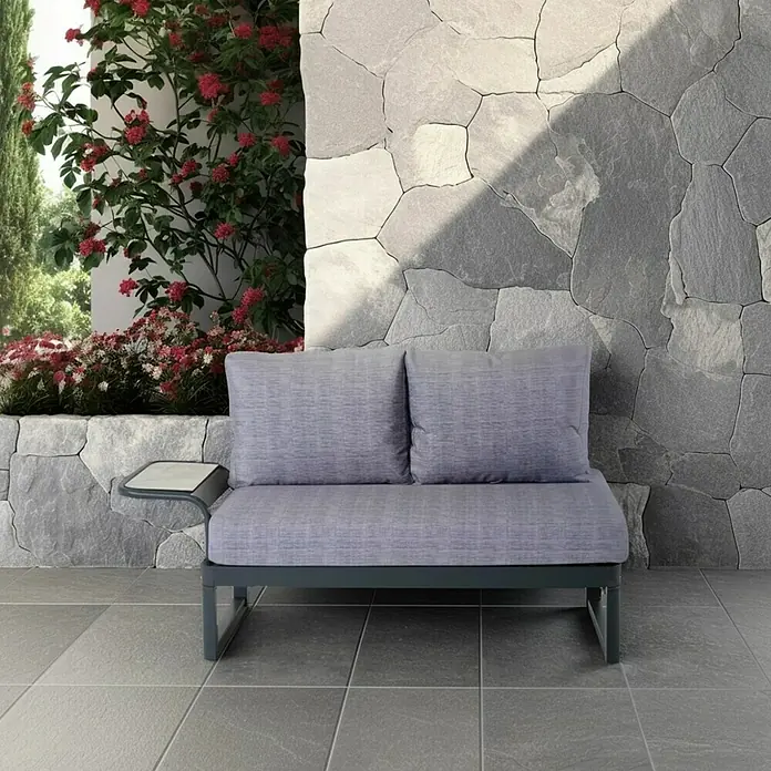 Bonlay Loungesofa Elara - B x T x H: 148 x 65 x 83 cm, Aluminium, Polyester, HPL, Anthrazit - Ein graues Gartensofa mit passenden Kissen steht vor einer Steinmauer, neben blühenden Pflanzen, und lädt zum Verweilen im Freien ein.