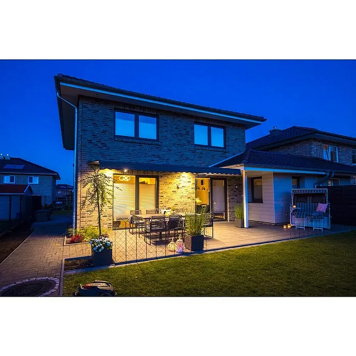 Spettmann Teilhülsenmarkise Sun LED 5822 - Beige, Farbe Gestell: Eisenglimmer matt, Breite: 5 m, Ausfall: 3 m - Modernes Backsteinhaus bei Dämmerung mit beleuchteter Terrasse, Gartenmöbeln und Rasenfläche.