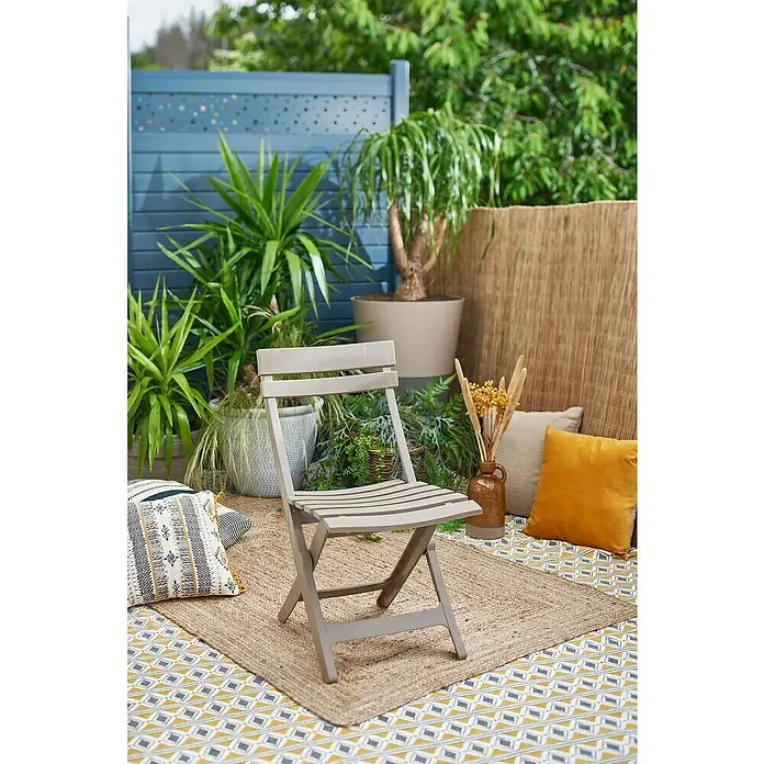 Grosfillex Klappstuhl Set Miami - 2 Stk., Polypropylen, Klappbar, Taupe, B x T x H: 42 x 50 x 93 cm - Ein beiger Klappstuhl steht auf einem geflochtenen Teppich auf einer Terrasse, umgeben von Pflanzen und Kissen.
