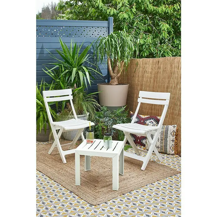 Grosfillex Garten-Beistelltisch Miami - L x B x H: 40 x 40 x 35 cm, Polypropylen, Stapelbar, Rechteckig, Weiß - Weißer klappbarer Outdoor-Tisch mit zwei Stühlen auf einer Terrasse mit Pflanzen, Getränken und Kissen.