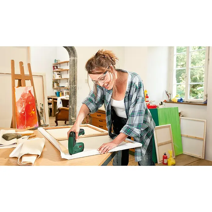 Bosch Akku-Tacker PTK 3,6 LI Office Set - 3,6 V, Klammertiefe: 4 mm - 10 mm - Frau benutzt einen Bosch-Tacker, um Leinwand auf einen Keilrahmen zu spannen. Künstleratelier mit Staffeleien und Gemälden im Hintergrund.
