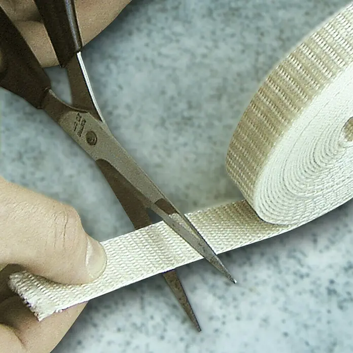 Schellenberg Cinta para persiana - Beige, Largo: 4,5 m, Anchura de la correa: 14 mm - Cortando un tejido de color claro con unas tijeras plateadas. El tejido está enrollado en un rollo.