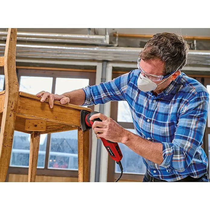 Einhell Expert Elektro-Deltaschleifer TC-DS 20 E - 200 W, 12.000 U/min - 22.000 U/min, Länge Schleifplatte: 90 mm - Mann schleift Holzstuhl mit einem roten Einhell-Multifunktionswerkzeug, Schutzbrille und Maske für die Sicherheit.