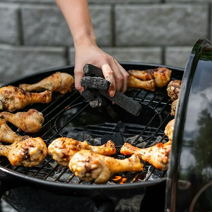 Char-Griller Holzkohlegrill - Durchmesser Grillfläche: 57 cm, Schwarz - Hand legt Holzkohle auf schwarzen Grill, goldbraune Hähnchenkeulen braten auf dem Rost.
