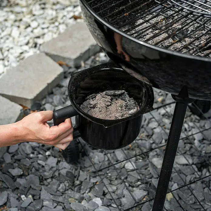 Char-Griller Holzkohlegrill - Durchmesser Grillfläche: 57 cm, Schwarz - Eine Hand hält einen schwarzen Holzkohle-Aschebehälter mit Asche und Kohlestücken neben einem Grill.