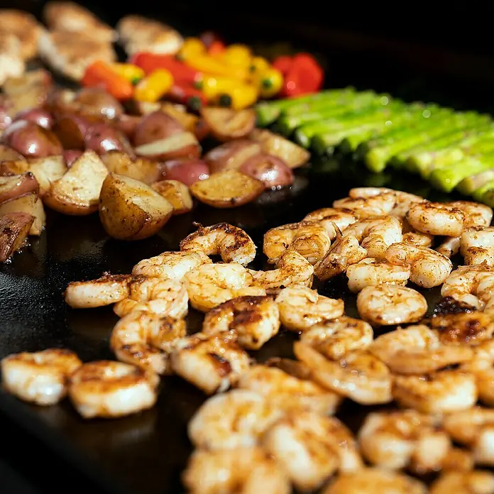 Char-Griller Plancha-Grill Flat Iron™ Premium Gas Griddle - Grillfläche (B x T): 56 x 46 cm, 14,65 kW, Anzahl Brenner: 4 Stk. - Garnelen, gewürzte Kartoffeln, bunte Paprika und grüner Spargel auf einer schwarzen Grillplatte.