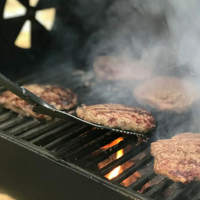 Char-Griller Smoker Portable Grill and Side Fire Box - Grillfläche (B x T): 45 x 43 cm - Hamburger brutzeln auf schwarzem Grill, einer wird mit Metallspatel in Rauch und Flammen gewendet.